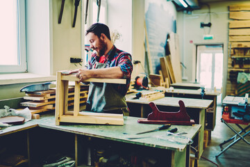 Wall Mural - bearded caucasian male artisan measuring handmade stool in own manufacture workshop business, skille