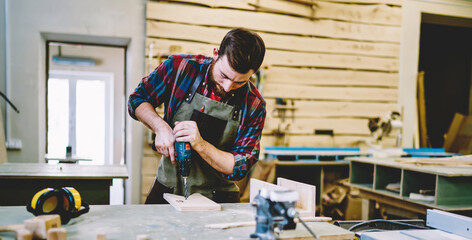 Wall Mural - skilled craftsman in apron using drill while working with wooden plank for furniture in own workshop
