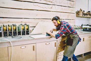 Wall Mural - serious male carpenter in headphones for protection using modern grinder during woodworks in worksho