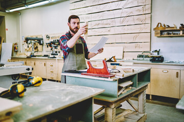 Wall Mural - thoughtful bearded male joiner holding wooden plank and paper with instruction for making furniture,