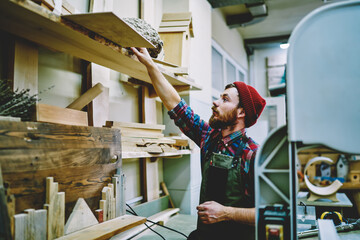 Wall Mural - caucasian bearded handyman in apron taking material from shelves in workshop for repairing wooden go
