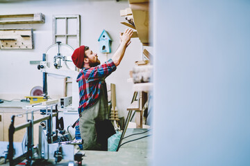 Young caucasian handyman taking stuff from top shelves in workshop making preparation for beginning work day, male carpenter looking for instruments and equipment in cabinetry in craft store.