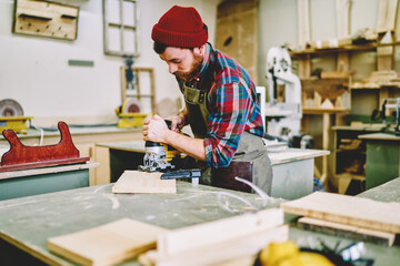 Wall Mural - serious bearded foreman in apron using modern electric machinery for woodenworks in workshop, skille
