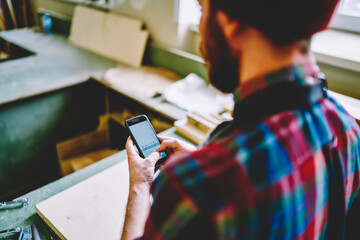 Cropped image of artisan using mobile phone during working day in workshop, back view of caucasian man holding smartphone and chatting in social networks with clients owning restoration service.