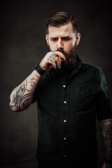 Wall Mural - Stylish bearded man smokes a cigar. Studio portrait of a handsome tattooed hipster guy who smokes a cigar