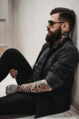 Wall Mural - Stylish bearded guy with tattoos on his hands and neck sitting on a floor in a white studio