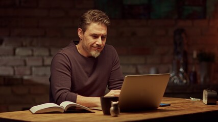 Older man tele working with laptop computer at home office in dark room, thinking, looking busy. 