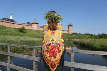 Summer in Russia. Russian tourist woman in traditional national Pavlovo Posad folk shawl, Pavloposadsky scarf. Flower wreath. Kremlin in Suzdal town, Vladimir region, Russia. Summer travel in Russia