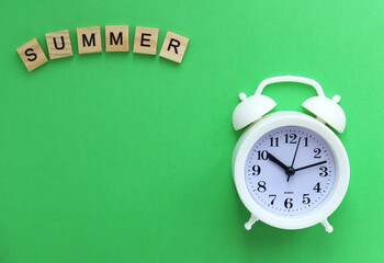 A white alarm clock and an inscription made of wooden plaques with letters: Summer, on a green background. Selective focus. Copy space