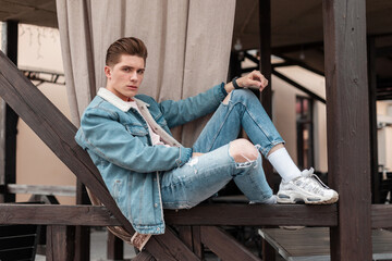 Good-looking young man in fashion casual blue jeans clothes in stylish white sneakers sit on wooden railing in city. Trendy fashionable guy in vintage youth denim wear in street. Summer menswear.