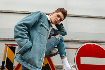 Wall Mural - Modern handsome young man in fashionable blue denim jacket in vintage jeans stands near metal road signs on street. Nice guy in trendy youth jeans clothes posing in city outdoors. Casual wear for men.