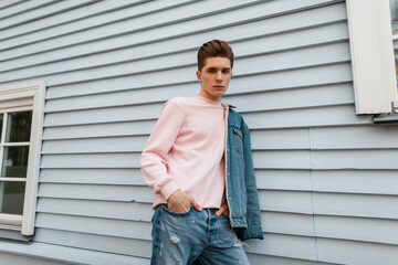 Wall Mural - Fashionable pretty young man in pink stylish sweatshirt in denim blue jacket in jeans stands near white vintage building on street in city. Trendy attractive guy fashion model with hairstyle outdoors.