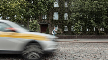 Wall Mural - Traffic concept. Cool young man tourist in trendy youth jeans clothes with fashion cloth bag travels on vintage city street near old building overgrown with green leaves. Urban guy walk near road.