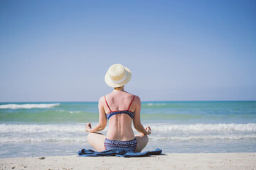 a girl in a swimsuit and a straw hat sits on the sand by the sea in a lotus lotus and meditates
