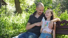A Father And A Little Girl Sit On A Bench In The Park And Talk. A Father Spends Time With His Young Daughter In The Park.