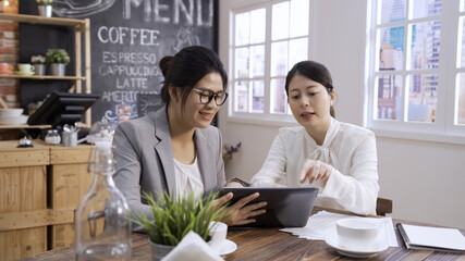 Wall Mural - Two confident asian japanese female businesspeople using digital tablet together while working at cafe table in modern cafe store. beautiful office lady coworkers talk and point on pad in cafeteria