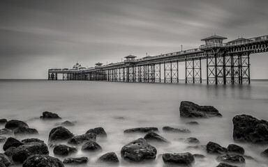 Long exposure black and white fine art photograph of Llandudno pier with rocks in the foreground. Smooth water and clouds in monochrome