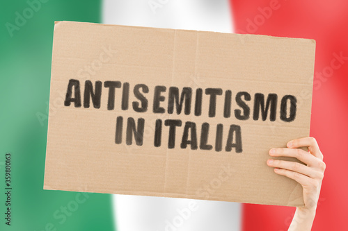Fotografía The phrase Antisemitism in Italy in Italian language on a banner in men's hand with blurred flag on the background