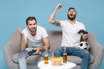 Displeased crazy guys friends sit on couch isolated on blue background. Sport leisure concept. Cheer up support favorite team with soccer ball play game with joystick screaming doing winner gesture.