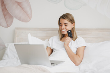 Beautiful young woman girl lying in bed with white sheet pillow blanket spending time in bedroom at home. Rest relax good mood lifestyle concept. Working on laptop pc computer hold credit bank card.