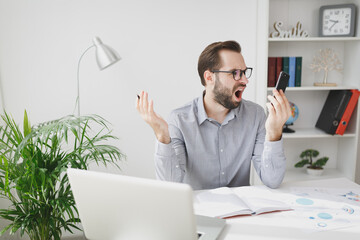 Wall Mural - Shocked irritated young business man in gray shirt sitting at desk working on laptop pc computer in light office on white wall background. Achievement business career concept. Talking on mobile phone.