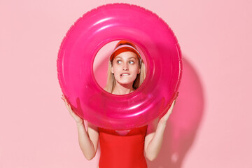 Wall Mural - Pensive young blonde woman girl in red one-piece swimsuit cap posing isolated on pink background. People summer vacation rest lifestyle concept. Hold swim inflatable ring, biting lips looking aside.