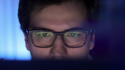 Man in eyeglasses late at night scrolling in front of laptop profile shot. Coder, programmer or developer using laptop in dark. Close up of glasses with reflection of computer screen.