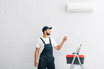 Wall Mural - Smiling workman switching air conditioner with remote controller near toolbox on ladder