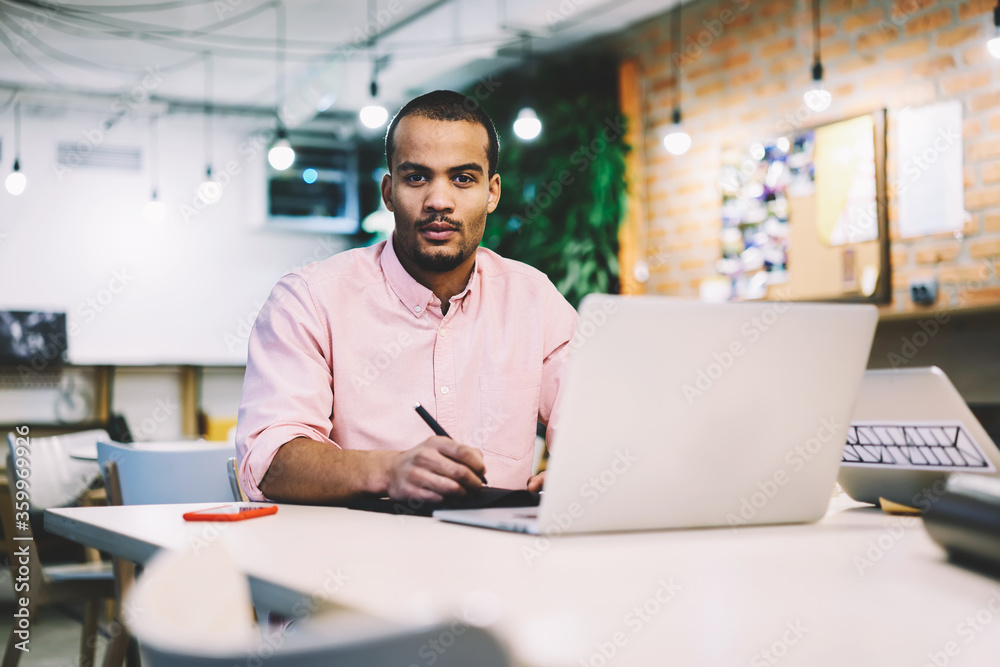 Portrait of concentrated male entrepreneur working with financial documentation and statistic reports in online database counting sum of costs and income of his business startup using laptop and wifi