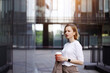 © Eva March - Business woman in beige suit, holding cup of coffee, walking outdoors on city street with skyscraper or building background, looking on window. Break at work, leisure time, enjoyment, shopping