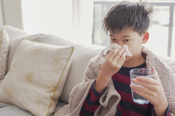 Mixed Asian sick preteen boy blowing nose and holding glass of water at home, heath care concept