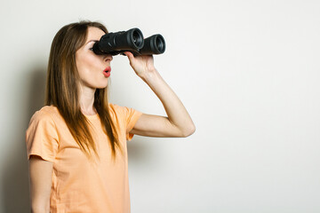 Wall Mural - Young friendly girl in a T-shirt looks through binoculars on a light background