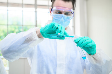 Male scientist wearing protection suit holding beaker or tube with sample liquid inside and working with many lab equipment for research at laboratory.