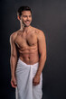 © Roman - Handsome young bearded man isolated. Topless muscular man in towel is standing on grey background. Serious man looking away.