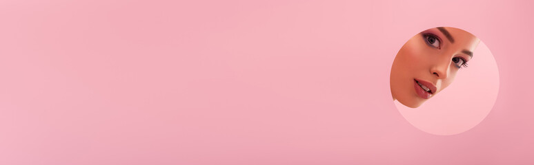 portrait of beautiful woman with shiny makeup in paper round hole isolated on pink, panoramic shot