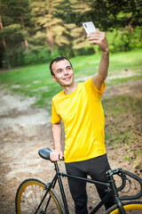 A young man taking a selfie with his Bicycle. In a public Park, among trees and vegetation.