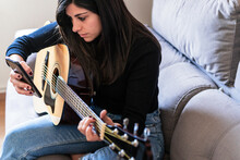Woman Playing Guitar Sitting On Her Couch At Home And Learning With Online Lessons And Some Masks Are Hanging Due To Containment. Behind It Is A Brick Wall