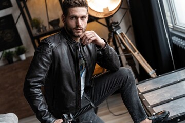 Wall Mural - Handsome young man in leather jacket looking away while sitting on the stool