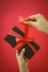 Girl holds a gift box and unties the bow. a girl opens a gift box. Gift box on a red background. Girl unties the tape from the box.
