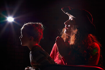 Wall Mural - Ugle oman in red sweater and pirate hat with curly hair posing with small boy on black background. Actress mother and actor son in magical, fantastic, fairy-tale movie in dark studio and flash