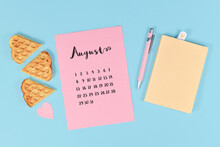 August Month Concept With Pink Calendar Sheet, Book, Pen, Paper Heart And Waffle Pieces On Ight Blue Background
