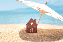 Miniature House And Umbrella On Beach, Blue Sea And Sky On Blurred Background. Vacation Home For Happy Holiday For Family. Real Estate, Sale Or Property Investment Concept. Closeup. Copy Space