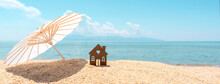 Small House And Umbrella On Sand, Beach, Blue Sea And Sky On Blurred Background. Real Estate, Sale Or Investment Property Concept. Symbol Of Dream Home For Family. Banner. Panoramic View. Copy Space