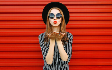 Wall Mural - Fashion portrait young woman blowing red lips sending sweet air kiss over red wall background