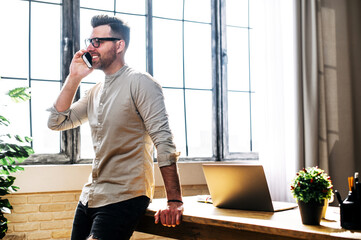 A guy in casual clothes and glasses is leaning on the table while talking over the phone, he stands in the light sunny office room, workplace with laptop on background, cosy office