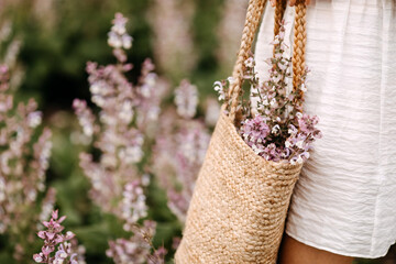 close-up of woman in white dress, standing in a big field with sage in bloom, holding a wicker bag w