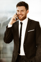 Business concept. Happy smiling young businessman standing in office talking on a cell phone getting good news about his work. Man in suit indoors on glass window background