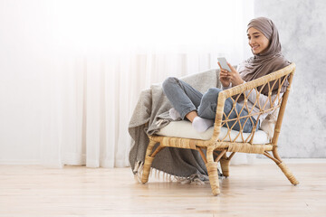 Canvas Print - Smiling Muslim Woman Sitting In Modern Chair With Smartphone, Relaxing At Home