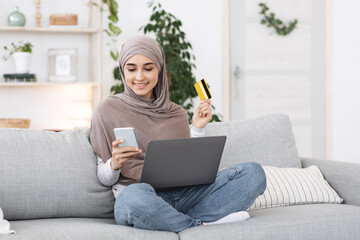 Wall Mural - Online Payments. Happy arabic girl using smartphone, laptop and card at home