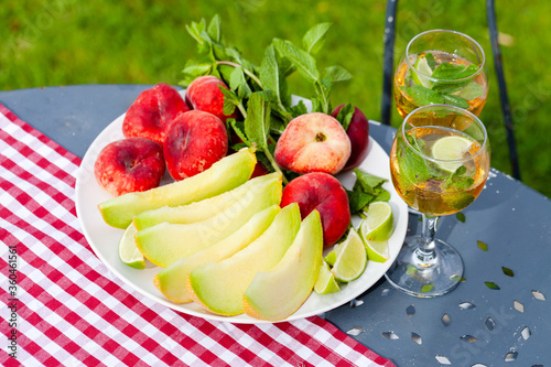 Summer party outdoor. Romantic picnic for a couple. Two glasses of cool wine served with mint and lime. Fresh melon and peach as simple light meal. Tasty low calories dessert. Green background.